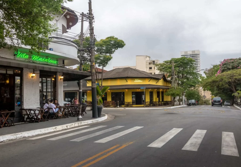 Imagem de uma rua tranquila na Vila Madalena, em São Paulo, mostra assentos ao ar livre em um café à esquerda, chamado Vila Madalena. Do outro lado da rua, há um prédio amarelo cercado por árvores. Poucas pessoas são vistas, e o céu está nublado