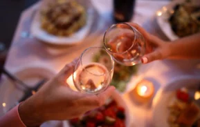 Imagem de duas pessoas batendo taças de vinho em uma mesa de jantar à luz de velas, com pratos de comida e uma pequena vela ao fundo, criando uma atmosfera calorosa e romântica