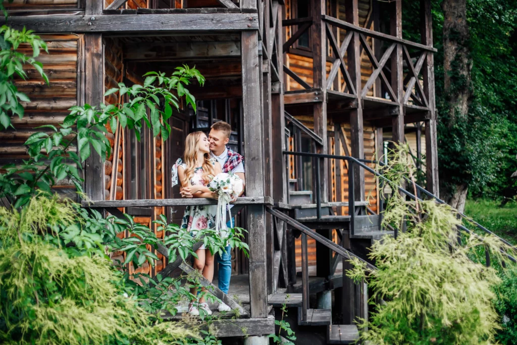 Imagem de um jovem casal na varanda de madeira de uma cabana rústica cercada por vegetação, sorrindo e se abraçando em um ambiente externo tranquilo