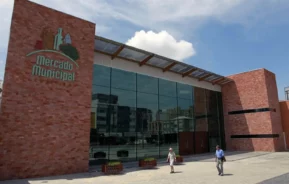 Imagem da estrutura externa do Mercado Municipal de Curitiba mostra um moderno edifício de tijolos. Duas pessoas caminham ao ar livre em uma praça pavimentada sob um céu parcialmente nublado