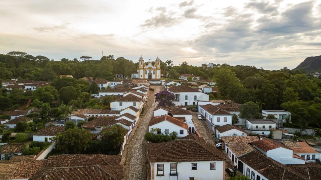 Imagem da vista aérea de Tiradentes, em Minas Gerais, mostra ruas de paralelepípedos, casas coloniais brancas com telhados de telha marrom e uma grande igreja amarela e branca em uma colina, cercada por vegetação sob um céu nublado