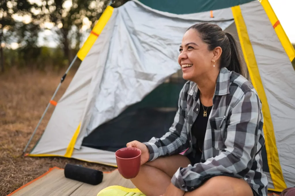 Imagem de uma mulher sentada em uma esteira em frente a uma barraca, sorrindo e segurando uma caneca vermelha. Ela usa uma camisa xadrez, shorts e tênis brilhantes, parecendo relaxada enquanto acampa ao ar livre. As árvores são visíveis ao fundo