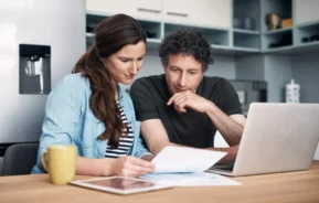 Imagem de um homem e uma mulher sentados em uma mesa de cozinha, revisando papéis juntos. Um notebook, um tablet e uma caneca amarela estão sobre a mesa. Ambos estão concentrados nos documentos. A cozinha é moderna, com armários de cor clara