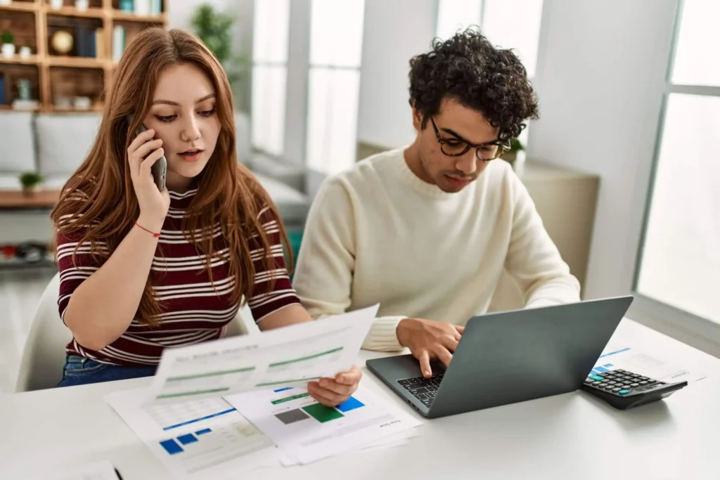 Imagem de dois jovens adultos trabalhando em uma mesa. A mulher fala ao telefone enquanto segura documentos, e o homem digita em um notebook. Os papéis e uma calculadora estão espalhados sobre a mesa