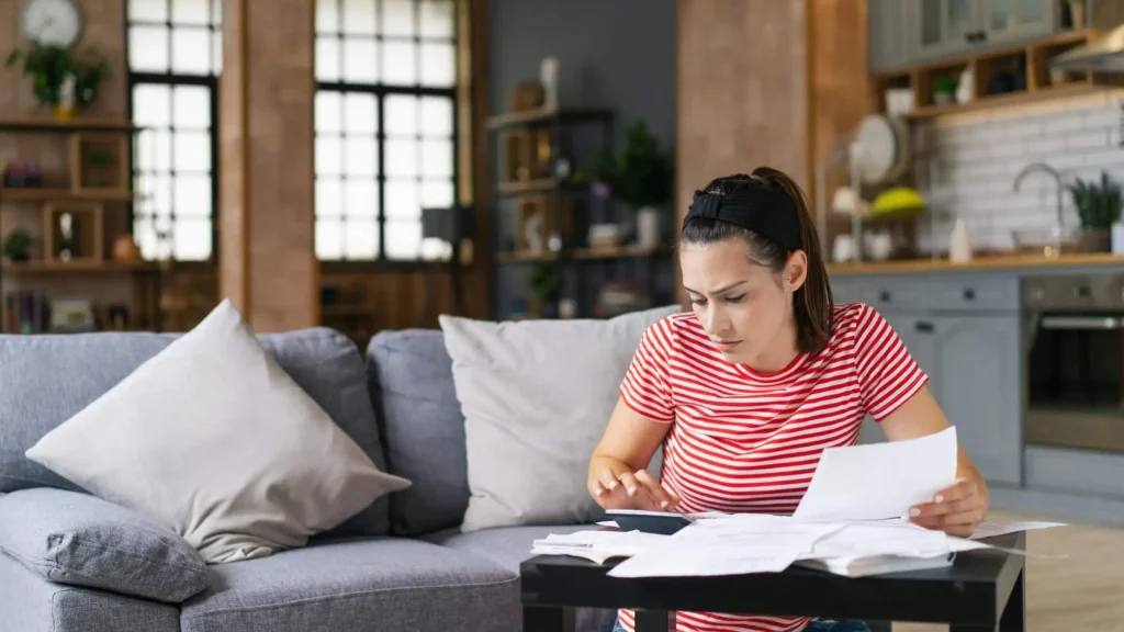 Imagem de uma mulher de camisa listrada vermelha sentada em um sofá na sala de estar, olhando papéis e usando uma calculadora, parecendo concentrada e preocupada. Uma mesa de centro com mais papéis está à sua frente