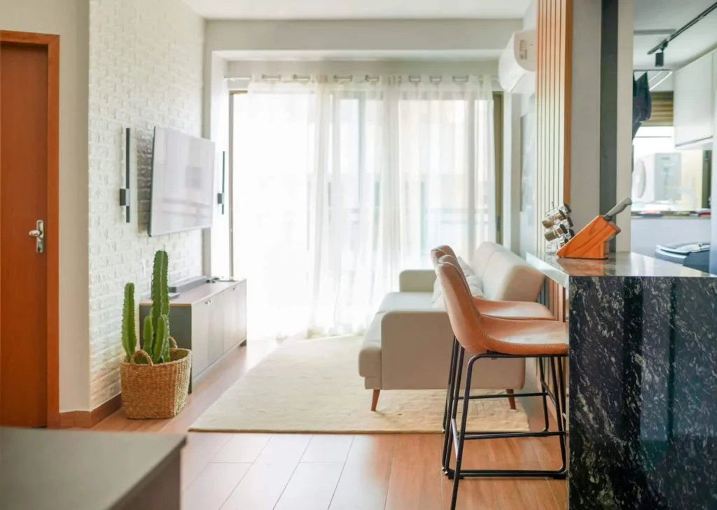 Imagem de uma sala de estar moderna e bem iluminada com um sofá cinza, TV na parede, uma cesta de tecido com um cacto, dois bancos de bar de couro em um balcão de mármore preto e cortinas transparentes que permitem a entrada de luz natural