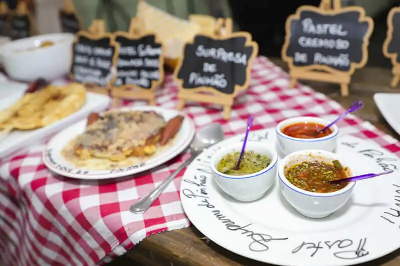 Imagem de três tigelas pequenas de salsa colorida com colheres em um prato branco, ao lado de outros pratos mexicanos, em uma toalha de mesa quadriculada vermelha e branca com placas de giz ao fundo