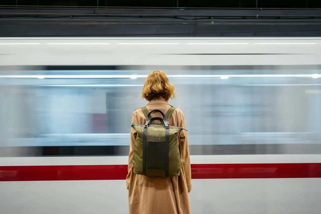 Imagem de uma pessoa de cabelo curto, vestindo um casaco bege e uma mochila verde, está em uma plataforma de metrô, de frente para um trem branco com uma faixa vermelha que passa. O trem está desfocado, indicando que está se movendo rapidamente
