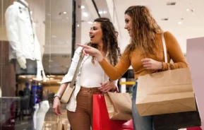 Imagem de duas mulheres jovens com sacolas de compras olhando e apontando para a vitrine de uma loja com uma jaqueta branca em um shopping center