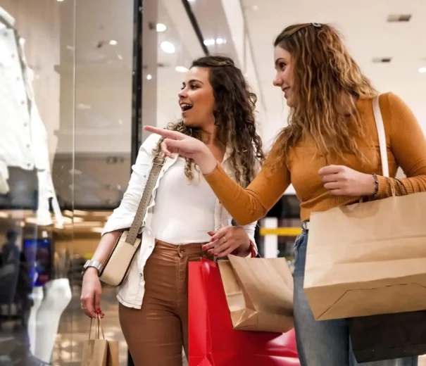 Imagem de duas mulheres jovens com sacolas de compras olhando e apontando para a vitrine de uma loja com uma jaqueta branca em um shopping center