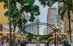 Uma animada rua de pedestres ladeada por árvores e edifícios altos, com um monumento de relógio no centro e pessoas caminhando em um dia nublado. Os postes de iluminação pública estão acesos ao longo do caminho.