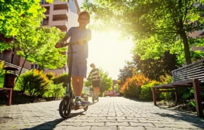 Um menino anda de scooter em um caminho iluminado pelo sol em um parque, sorrindo para a câmera. Outra criança anda de scooter ao fundo, cercada por árvores e prédios em um condomínio verde.
