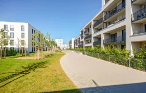 Prédios de apartamentos modernos com varandas, parte de uma instituição de condomínio, alinham-se a uma passarela pavimentada cercada por gramados verdes e árvores jovens, sob um céu azul claro em um dia ensolarado.