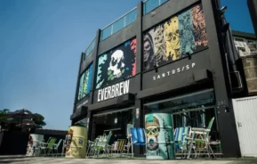 Foto que ilustra matéria sobre os melhores bares em Santos mostra a fachada do Everpub, com mesas ao ar livre e barris decorados na frente, sob um céu azul claro (Foto: Divulgação)