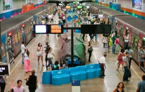 Uma plataforma de estação de metrô movimentada com muitas pessoas andando, em pé e esperando. Dois trens estão parados em cada lado, e assentos e placas coloridas são visíveis em toda a área bem iluminada.