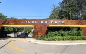 Entrada do Parque do Paço, com uma fachada moderna de madeira com letras brancas em negrito, cercada por vegetação e localizada ao lado de uma estrada pavimentada com marcações amarelas. O céu azul e as árvores são visíveis ao fundo.