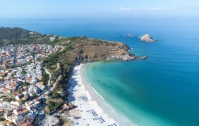 Vista aérea de uma cidade costeira com prédios brancos agrupados perto de uma praia de Arraial do Cabo, na Região dos Lagos do Rio de Janeiro, de areia, água azul-turquesa e um promontório rochoso que se estende até o mar sob um céu azul claro. Os guarda-sóis estão dispostos ao longo da costa.