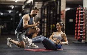 Imagem de um personal trainer ajoelhado ao lado de uma mulher sorridente segurando um kettlebell, enquanto ela se exercita em uma esteira em uma academia, com pesos e equipamentos de ginástica ao fundo
