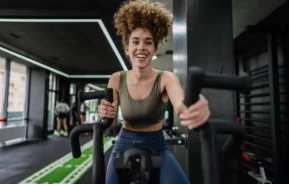 Imagem de uma mulher de cabelos cacheados sorrindo enquanto usa uma bicicleta ergométrica em uma academia, vestindo um sutiã esportivo verde e leggings azuis. Outros equipamentos de ginástica e pessoas são visíveis ao fundo