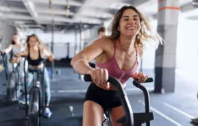 Imagem de uma mulher sorridente usando um top esportivo rosa em uma bicicleta ergométrica de uma academia, com outras mulheres se exercitando em bicicletas ao fundo. O cenário é brilhante e energético
