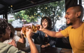 Imagem de quatro amigos sentados ao redor de uma mesa em um bar ao ar livre, sorrindo e rindo enquanto batem seus copos de cerveja em um brinde. Luzes de corda estão penduradas acima deles e árvores podem ser vistas do lado de fora