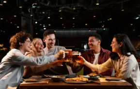 Imagem de cinco amigos sentados ao redor de uma mesa de restaurante, sorrindo e levantando copos de cerveja em um brinde, com pratos de comida e pizza à sua frente, em um ambiente moderno e bem iluminado