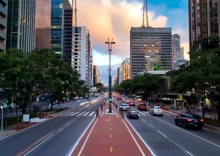 Imagem da Avenida Paulista mostra rua larga com prédios altos e modernos, árvores ao longo das calçadas, carros em ambas as direções e um céu colorido com nuvens ao pôr do sol. Uma ciclovia vermelha passa pelo centro
