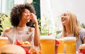 Imagem de duas mulheres sentadas em uma mesa ao ar livre, rindo e saboreando alimentos e bebidas. Há hambúrgueres, batatas fritas e canecas de cerveja na frente delas. A atmosfera é clara e alegre