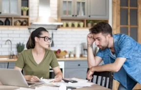 Imagem de um homem e uma mulher sentados à mesa da cozinha coberta de papéis, um notebook e uma calculadora. A mulher olha para o homem, que parece estressado, enquanto eles analisam documentos juntos