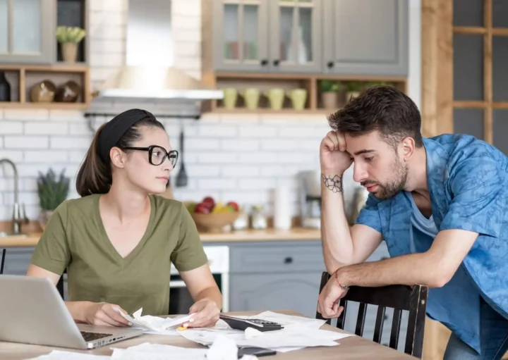 Imagem de um homem e uma mulher sentados à mesa da cozinha coberta de papéis, um notebook e uma calculadora. A mulher olha para o homem, que parece estressado, enquanto eles analisam documentos juntos