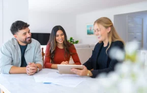 Imagem de um casal sorridente sentado à mesa com uma mulher profissional que lhes mostra algo em um tablet, com papéis espalhados sobre a mesa em uma casa moderna e bem iluminada