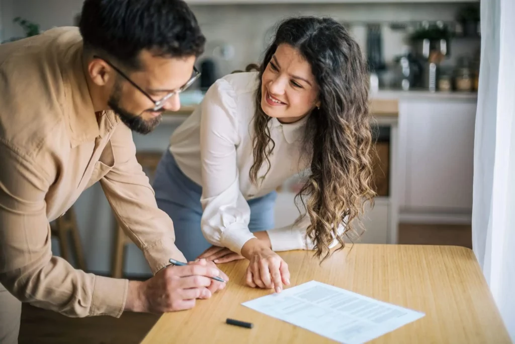 Imagem de um homem assinando um documento em uma mesa de madeira enquanto uma mulher ao seu lado sorri e aponta para o papel. Eles parecem estar em uma cozinha bem iluminada