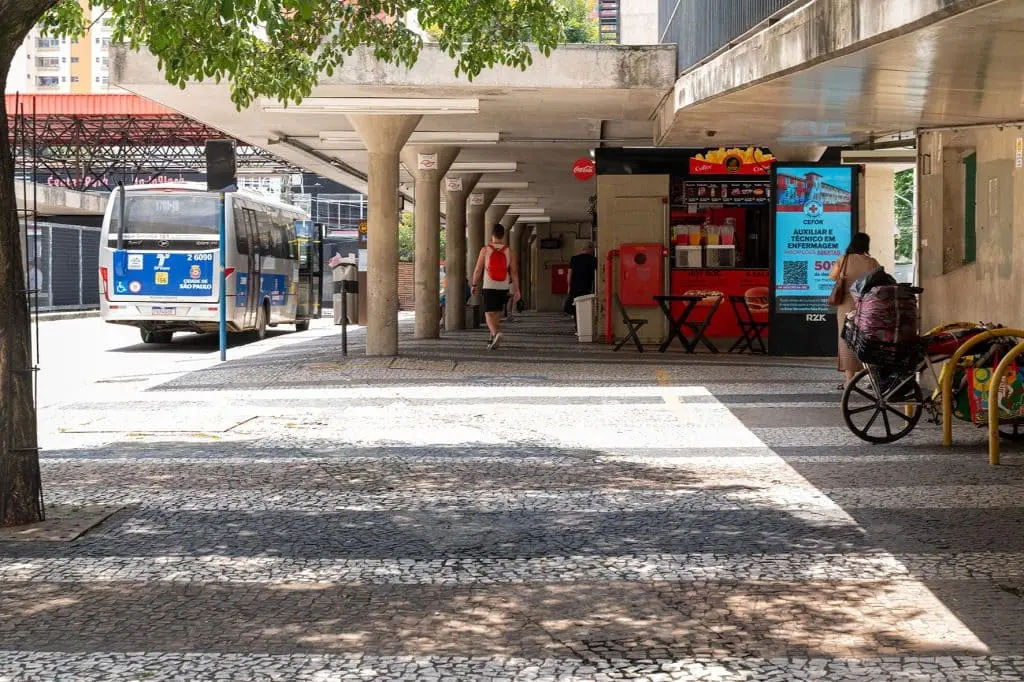 Imagem do terminal da estação Parada Inglesa, em São Paulo, em um dia ensolarado, mostra um ônibus azul estacionado à esquerda, pessoas caminhando, uma mulher com uma bicicleta à direita e uma lanchonete com cadeiras e mesas vermelhas sob um prédio de concreto
