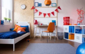 Imagem de um quarto de menino moderno e arrumado com uma cama de solteiro, detalhes em azul e laranja, uma escrivaninha com uma cadeira laranja, prateleiras com caixas e livros, decorações de parede e luz natural de uma janela