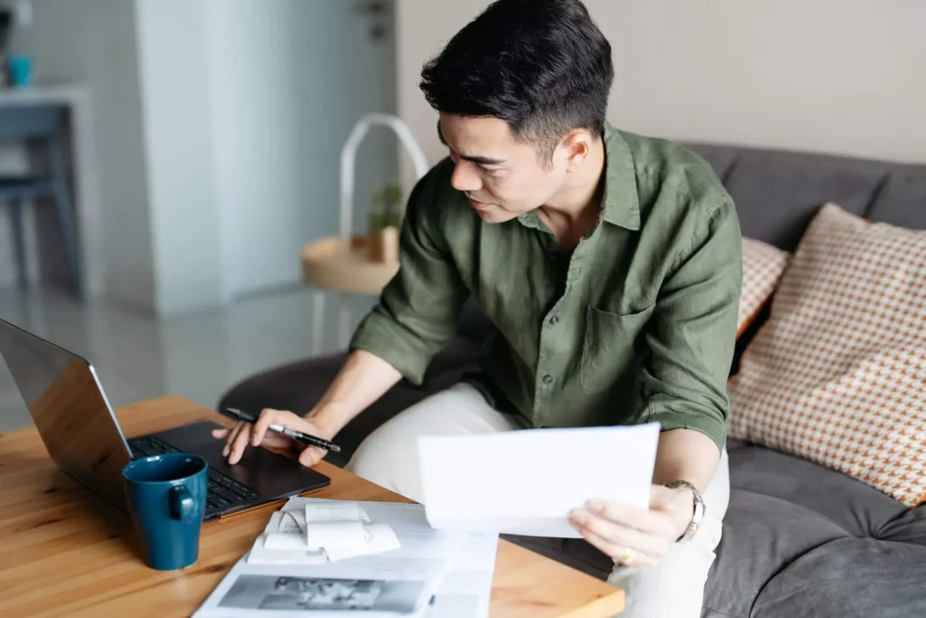 Imagem de um homem sentado em um sofá, trabalhando em uma mesa de madeira com um laptop, segurando papéis e uma caneta. Há uma caneca azul e mais documentos sobre a mesa. Ele está concentrado em seu trabalho em um ambiente doméstico