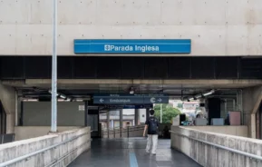 Imagem de uma pessoa caminhando em direção à entrada da estação de metrô Parada Inglesa, em São Paulo, com uma placa azul da estação acima e placas direcionais para embarque e bilhetes visíveis no interior