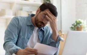 Imagem de um homem usando óculos e camisa jeans sentado em uma mesa com um laptop, segurando uma folha de papel e apoiando a testa na mão, parecendo estressado ou preocupado