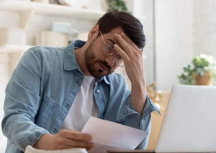 Imagem de um homem usando óculos e camisa jeans sentado em uma mesa com um laptop, segurando uma folha de papel e apoiando a testa na mão, parecendo estressado ou preocupado