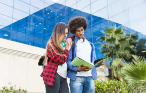 Imagem de dois estudantes do lado de fora de um moderno edifício de vidro, olhando juntos para um notebook. Um deles usa camisa xadrez vermelha e fones de ouvido, o outro tem cabelos cacheados e óculos. Há palmeiras e vegetação ao fundo