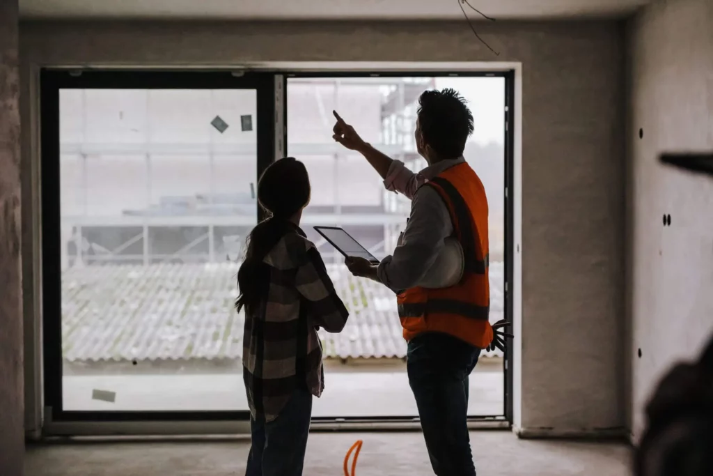 Imagem de uma pessoa com colete de segurança aponta para uma grande janela enquanto segura um laptop, ao lado de outra pessoa dentro de um prédio inacabado