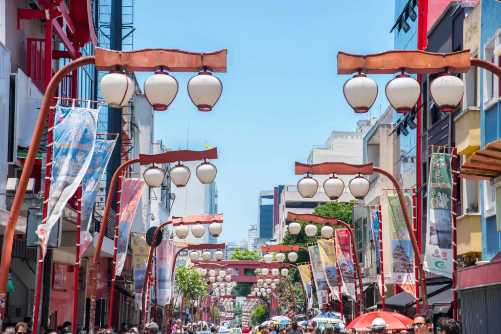 Imagem de uma rua do bairro Liberdade, em São Paulo, decorada com lanternas em estilo japonês penduradas em arcos vermelhos, cercada por prédios altos e faixas coloridas em um dia ensolarado