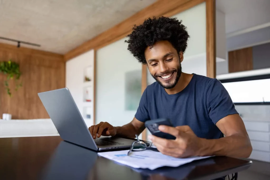 Imagem de um homem sorridente sentado em uma mesa com um laptop, segurando um smartphone em uma das mãos e olhando para ele. Papéis e óculos estão sobre a mesa à sua frente em um ambiente doméstico moderno