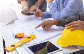 Imagem de três pessoas discutindo plantas arquitetônicas em uma mesa coberta com ferramentas de construção, um capacete amarelo, um tablet, uma fita métrica e um nível de bolha de ar. Uma pessoa aponta para as plantas enquanto outra segura um telefone
