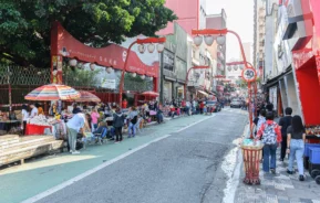 Imagem de avenida no bairro Liberdade, em São Paulo, mostra lanternas vermelhas penduradas acima e prédios nos dois lados da rua e pessoas caminhando pela calçada