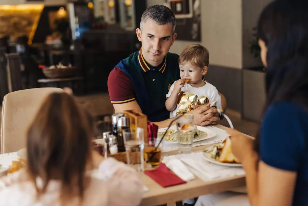 Imagem de um homem segurando uma criança pequena no colo em uma mesa de restaurante, sentado com uma mulher e uma menina. Eles estão comendo e bebendo juntos em um ambiente de jantar casual