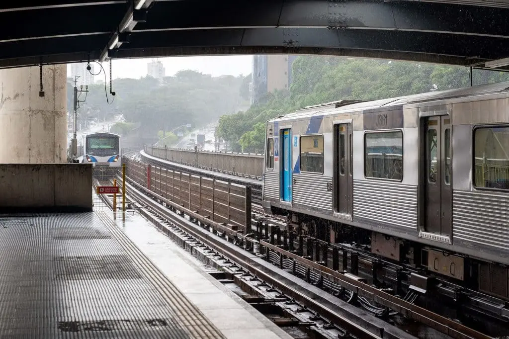 Imagem de um trem prateado esperando em uma plataforma externa da estação de metrô Parada Inglesa, em São Paulo, em um dia chuvoso, com outro trem se aproximando à distância. Trilhos molhados e árvores enevoadas são visíveis ao fundo