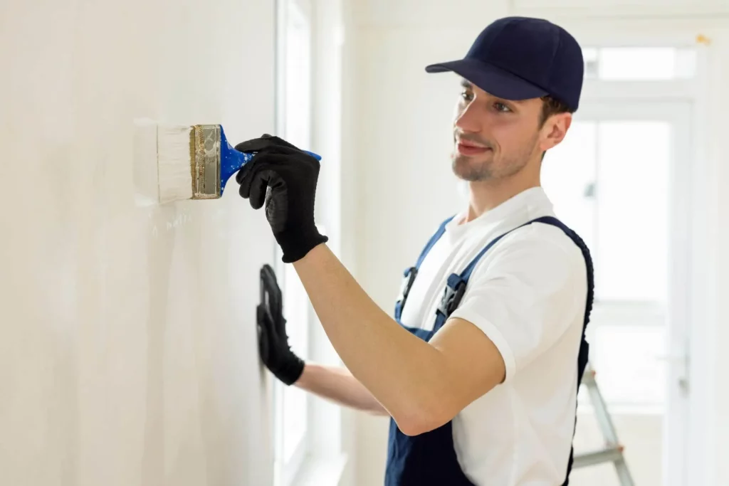 Imagem de um homem usando um boné azul-marinho, luvas pretas e macacão usa um pincel para pintar uma parede branca em um ambiente interno. A luz natural entra pelas janelas próximas