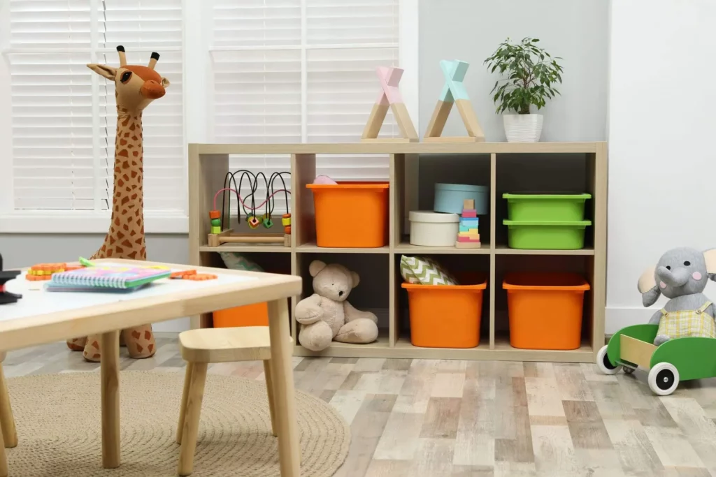 Imagem de um quarto de menino com uma prateleira com nichos laranja e verde, brinquedos e livros; uma girafa de pelúcia, um ursinho de pelúcia e um elefante; uma pequena mesa com cadeiras e uma planta na prateleira. Piso de madeira clara e persianas brancas