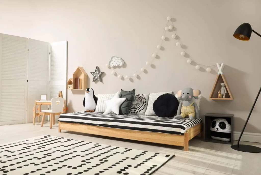 Imagem de quarto montessoriano com uma cama baixa de madeira, roupa de cama preta e branca, brinquedos de pelúcia de animais, decoração geométrica na parede, uma pequena escrivaninha com cadeira, luzes de corda, uma luminária de chão e um tapete de bolinhas em um piso claro