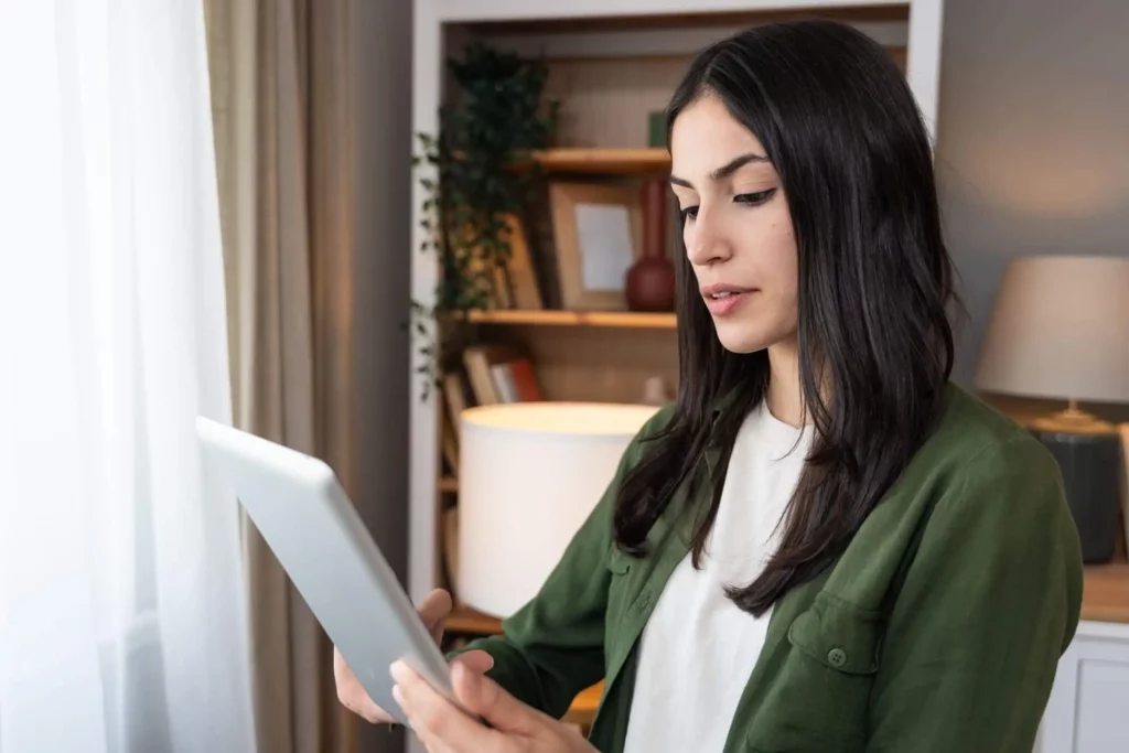 Imagem de uma mulher com longos cabelos escuros, vestindo uma jaqueta verde e camisa branca, olhando para um tablet em uma sala iluminada com estantes e uma lâmpada ao fundo